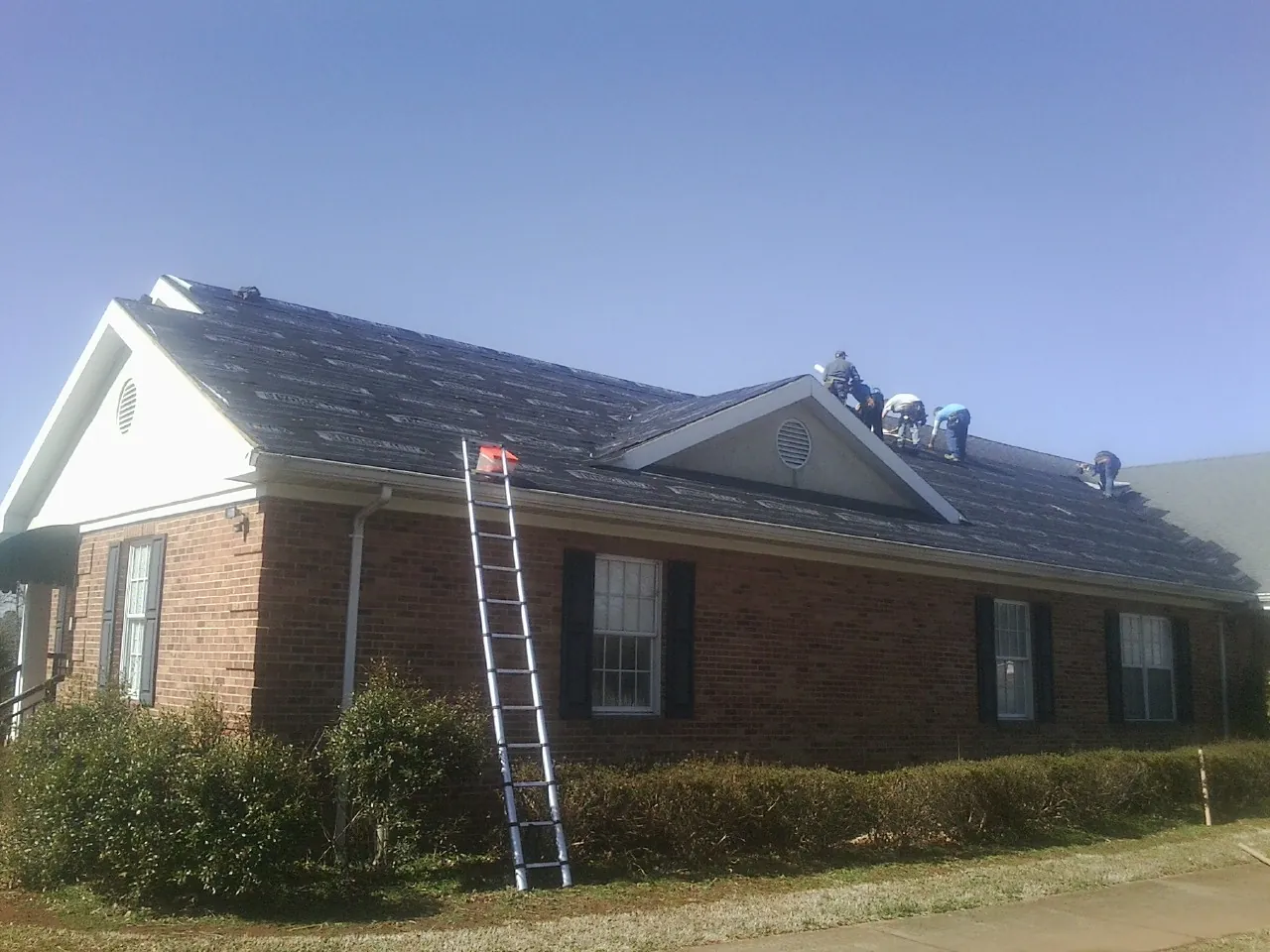 Crew tearing shingles off a brick ranch roof with a ladder set — Emergency Roof Repair work by our crew in Eastover