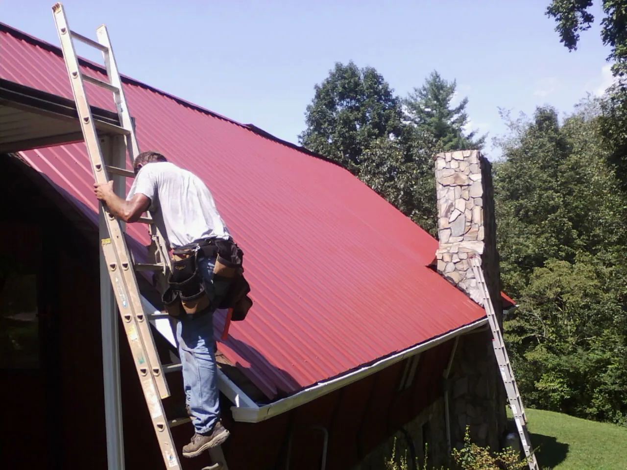 Roofer on a ladder beside a red metal roof and stone chimney — Soffit & Fascia Repair by Roofing Brothers in Eastover
